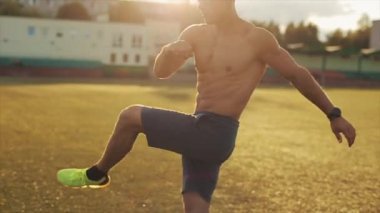 Spor şortlu bir vücut geliştirici stadyumda antrenman yapıyor ve gün batımında bacaklarını sallıyor. Sağlıklı bir yaşam tarzı. Yan görüş. Yakın plan.