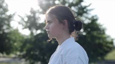 Beyaz kimonolu bir sporcu kadın doğanın arka planına karşı duruyor ve kameraya doğru karate duruşunu sergiliyor. Yakın plan. Ağır çekim. Kamera uzaklaşıyor.