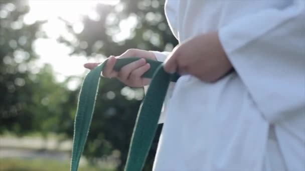 空手の女の子選手は白い着物で自然の中に立って、緑のベルトを結びます。手を閉じる。スローモーション。ぼやけた背景 — ストック動画