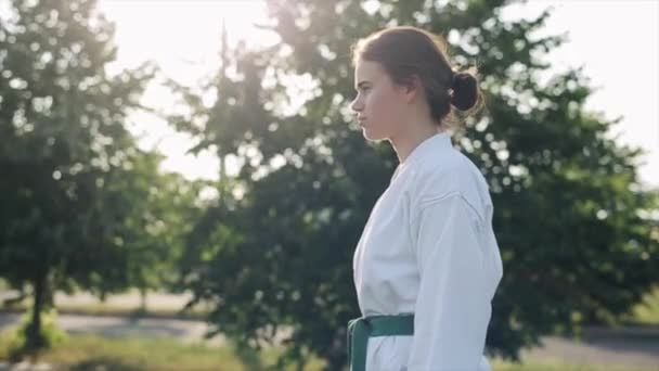 Egy kimonós fiatal nő a természetben edz, és karate standot mutat. Közelkép. Lassú mozgás. Homályos háttér — Stock videók