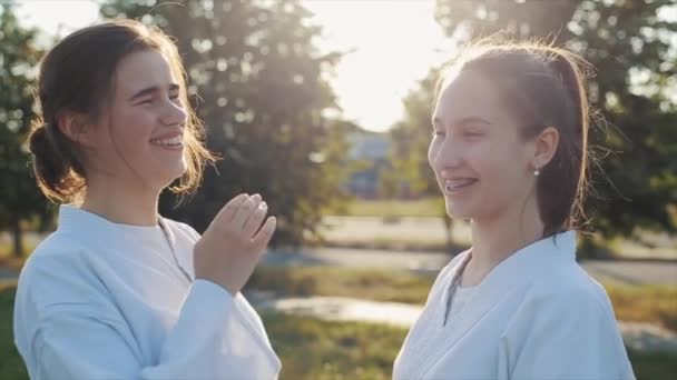 Junge Mädchen in weißen Kimonos stehen sich gegenüber und lachen, während sie im Freien Karate üben. Seitenansicht. Nahaufnahme. Zeitlupe — Stockvideo