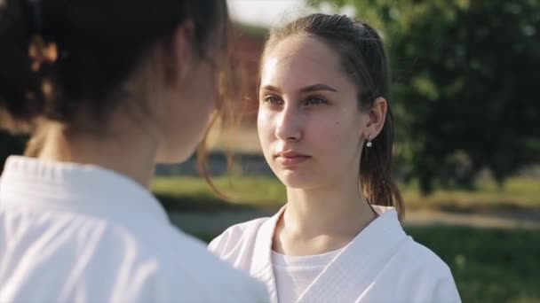 Młoda kobieta w białym kimono nawiązuje kontakt wzrokowy ze swoim partnerem karate podczas treningu na świeżym powietrzu. Zbliżenie. Zwolnij trochę. Skup się na twarzy sportowców. — Wideo stockowe