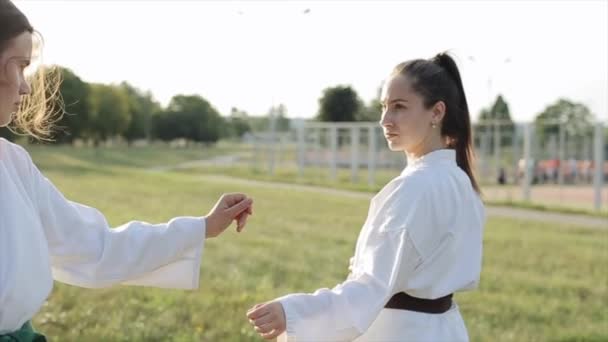 Två unga flickor står i en karate stå mot varandra mot bakgrund av en idrottsplats och träd. Närbild. Sakta i backarna. suddig bakgrund — Stockvideo