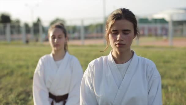 Két fiatal lány fehér kimonóban ül a fűben, és csukott szemmel meditálnak a karate edzés előtt. Előre nézz. Közelkép. Homályos háttér — Stock videók