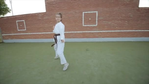 A fiatal lány fehér kimonóban harcművészeti technikákat gyakorol a karate edzés alatt a sportpályán. Előtér. Közelkép — Stock videók