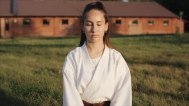 Karate eğitimi alan genç bir kadın çimenlerin üzerinde gözleri kapalı oturup meditasyon yapıyor ve gözlerini açıyor. Ön manzara. Yakın plan. Ağır çekim. Kamera yakınlaştır