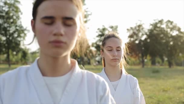 Zwei Karate-Mädchen führen eine Entspannungs- und Konzentrationseinheit in der Natur durch. Frontansicht. Nahaufnahme. Zeitlupe. Die Kamera zoomt heraus und ändert den Fokus — Stockvideo