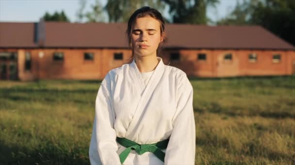 Chica de karate joven en un kimono blanco y un cinturón verde medita sentado en la hierba en la naturaleza. Vista frontal. Primer plano. En cámara lenta. Fondo borroso — Vídeos de Stock