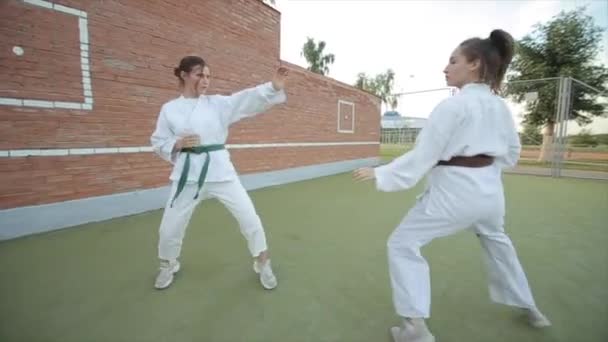 As mulheres jovens em quimonos brancos têm uma luta de sparring no treinamento de karatê em um campo de esportes. Close-up. A câmera entra e sai e se move em torno do assunto — Vídeo de Stock