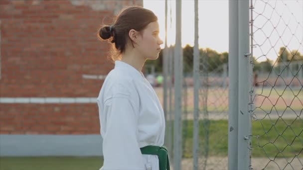 Beyaz kimonolu genç bir kız tuğla bir duvarın arkasında dururken karate standı gösteriyor. Ön manzara. Yakın plan. Yavaş çekim — Stok video
