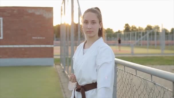 Egy fiatal lány egy fehér kimonóban a kiképzés bemutatja a különböző harci karate álláspontok, míg a sportpályán. Előre nézz. Közelkép — Stock videók