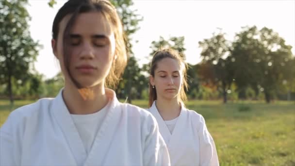 Karate sporcusu kadınlar doğada meditasyon yaparlar ve kızlardan biri gözlerini açar ve kameraya bakar. Yakın plan. Ağır çekim. Kamera uzaklaşıyor ve odağı değiştiriyor. — Stok video