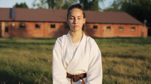 Una hermosa joven en clases de karate se dedica a la meditación sentada en la hierba. Vista frontal. Primer plano. En cámara lenta. Fondo borroso — Vídeo de stock