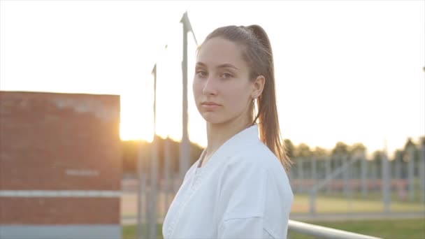 Retrato de una hermosa joven en un kimono blanco que muestra técnicas de karate en el campo de deportes sobre el fondo de la puesta del sol. Movimiento lento — Vídeos de Stock