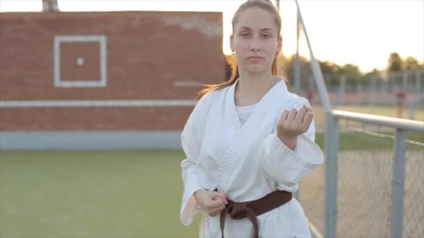 Egy fiatal nő fehér kimonóban edz a sportpályán, és bemutatja az alapvető karate pózokat. Közelkép. Lassú mozgás. Homályos háttér — Stock videók