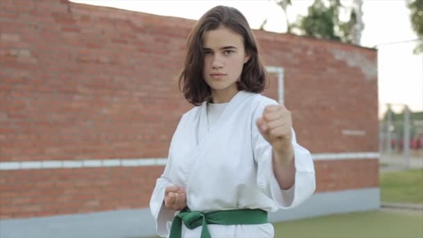Beyaz kimonolu genç bir kadın spor sahasında temel karate duruşunda duruyor ve kameraya bakıyor. Ön manzara. Yakın plan. Kamera sağa sola hareket ediyor. — Stok video