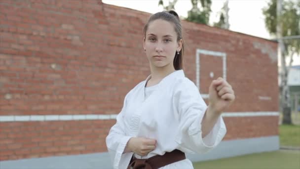 Egy fiatal lány portréja fehér kimonóban, aki karate alapállásba áll egy téglafal hátterében. Lassú mozgás. A kamera egyik oldalról a másikra mozog — Stock videók