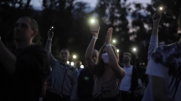 Misk, Biélorussie - 25 août 2020 : Une jeune fille portant un masque de protection se tient parmi de nombreuses personnes lors d'un événement ou d'un concert de masse et applaudit en tenant un téléphone portable avec une lampe de poche allumée. Gros plan — Video