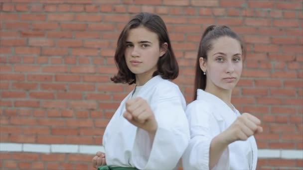 Junge Sportlerinnen in weißem Kimono stehen in Karate-Kampfpositionen vor dem Hintergrund einer Ziegelwand. Nahaufnahme. Zeitlupe. Die Kamera bewegt sich von links nach rechts und zurück — Stockvideo