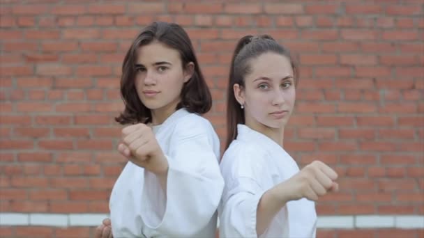 Twee jonge meisjes in witte kimono 's staan met hun rug naar elkaar toe en demonstreren een beschermende houding in karate tegen een bakstenen muur. Een close-up. De camera zoomt in en uit. Langzame beweging — Stockvideo