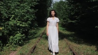 Beyaz elbiseli güzel bir genç kız bir yaz günü ormanda terk edilmiş eski bir demiryolu boyunca yürür. Ön manzara. Ağır çekimde yakın çekim