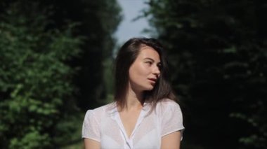 Güneşli bir yaz gününde yeşil ağaçların arka planında yürüyen beyaz elbiseli hoş bir genç kızın portresi. Yavaş çekim
