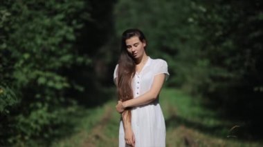 Gülümseyen genç bir kız, arka planda duran yeşil ağaçların önünde uzun saçlarını eliyle düzleştiriyor. Ön manzara. Yakın plan. Bulanık arkaplan