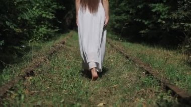 Beyaz elbiseli ve uzun saçlı genç bir kadın demiryolunun eski paslı rayları arasındaki çimlerin üzerinde yalınayak yürüyor. Arkadan bak. Yakın plan. Kamera aşağıdan yukarıya hareket eder