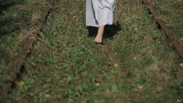 Une femme vêtue d'une longue robe blanche marche pieds nus sur l'herbe au milieu d'un vieux chemin de fer abandonné. Vue de face. Gros plan sur les jambes. Mouvement lent — Video
