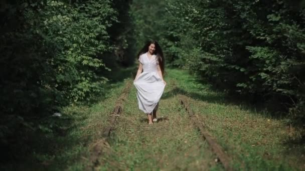 Uma jovem mulher sorridente com cabelos longos corre descalça entre os trilhos enferrujados de uma ferrovia abandonada na floresta segurando seu vestido com a mão. Vista frontal. Movimento lento — Vídeo de Stock