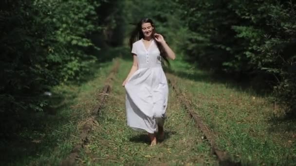 Привлекательная молодая женщина с трепещущими волосами бегает босиком среди зеленых деревьев в белом длинном платье. Вид спереди. Крупный план. Медленное движение — стоковое видео