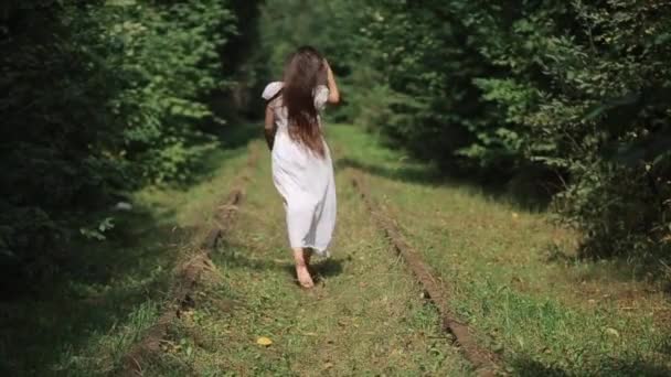 Een jong meisje in een witte jurk loopt blootsvoets langs het spoor van een oude verlaten spoorweg in het bos, recht haar lange haar met haar hand. Achteraanzicht. Langzame beweging — Stockvideo