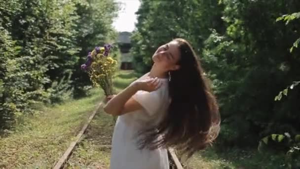 Una atractiva joven con el pelo revoloteando en el viento se levanta con un ramo de flores silvestres entre los árboles verdes. Vista lateral. Primer plano. En cámara lenta. La cámara cambia de enfoque — Vídeos de Stock