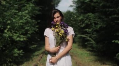 Yumuşak bir genç kadın elinde bir buket kır çiçeğiyle ormanın ortasında duruyor ve elleriyle saçlarını düzeltirken gülümsüyor. Ön manzara. Yakın plan.
