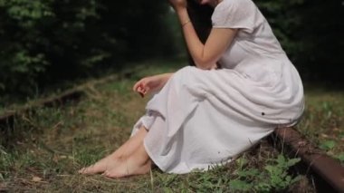 Romantik genç bir kadın terk edilmiş bir demiryolunun raylarında yeşil ağaçların önünde yalınayak oturuyor. Yan görüş. Yakın plan. Kamera aşağıdan yukarıya hareket eder