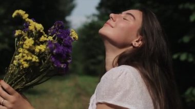 Beyaz elbiseli hassas genç bir kadın bir buket kır çiçeğiyle doğanın arka planında oturur ve onları koklayarak gözlerini kapatır. Yakın plan. Ağır çekim. Yan görünüm