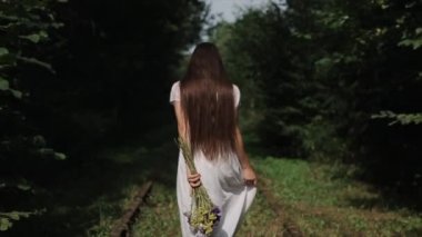 Beyaz elbiseli hoş bir genç kadın elinde bir buket çiçekle yeşil ağaçların arasında yürür ve kameraya bakar. Arkadan bak. Yavaş çekim