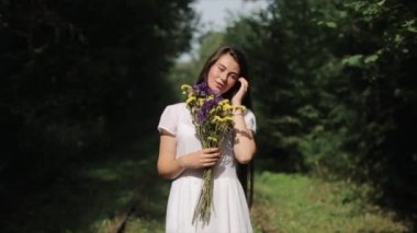 Beyaz elbiseli güzel bir genç kadın bir buket kır çiçeğiyle doğanın arka planına karşı duruyor ve elleriyle saçlarını düzleştiren kameraya poz veriyor. Manzara. Öne geç. Yakın plan.