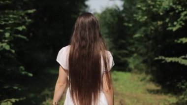 Genç uzun saçlı bir kız ağaçlar arasında bir patika boyunca yürür ve kameraya döner havaya çiçekler atar. Arkadan bak. Yakın plan. Yavaş çekim