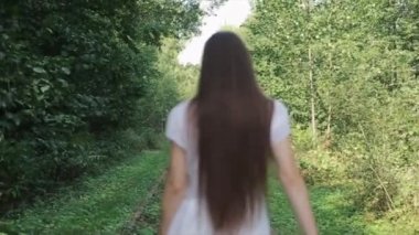 Beyaz elbiseli şehvetli genç bir kadın ağaçların arasında bir patika boyunca yürüyor ve kameraya dönüp havaya çiçekler atıyor. Yakın plan. Ağır çekim. Kamera odağı değiştirir.