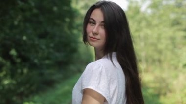 Hoş, gülümseyen genç bir kız dönüp kameraya bakar ve ağaçların arasında ilerlemeye devam eder. Yakın plan. Arkadan bak. Yavaş çekim