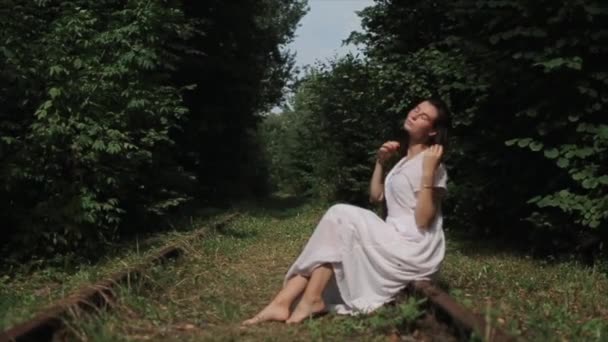 Una sensual joven con un vestido blanco se sienta entre los árboles verdes y endereza su largo cabello con las manos exponiendo su rostro al sol. Vista lateral. Primer plano — Vídeo de stock