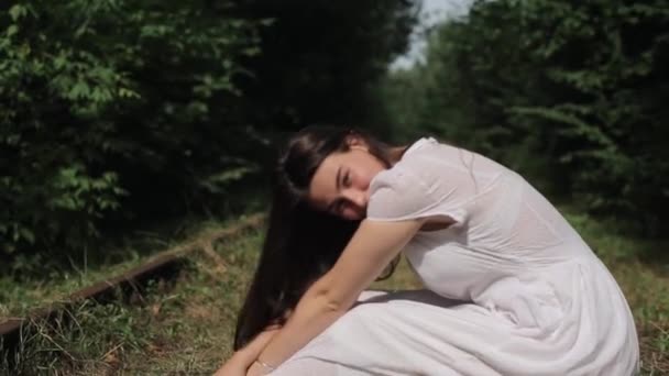 En vacker ung flicka i vit klänning sitter på de gamla rostiga rälsen på en övergiven järnväg i skogen och ler med huvudet lutat. Sidovy. Närbild. Kameran rör sig uppifrån och ner — Stockvideo