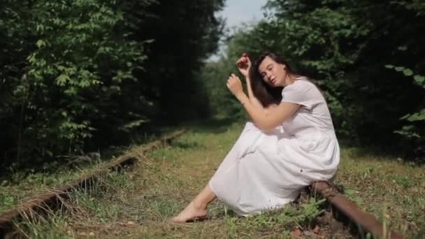 Μια εντυπωσιακή νεαρή γυναίκα με λευκό φόρεμα κάθεται ξυπόλητη στις σκουριασμένες ράγες ενός παλιού σιδηροδρόμου στο δάσος με το κεφάλι της λυγισμένο στα γόνατα. Πλευρική άποψη. Κοντινό πλάνο — Αρχείο Βίντεο