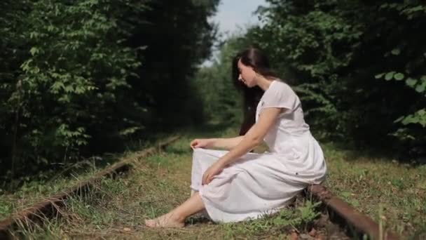 En blygsam ung flicka sitter barfota på spåren av en gammal övergiven järnväg i skogen och lutar huvudet räta ut sin långa vita klänning. Sidovy. Närbild — Stockvideo
