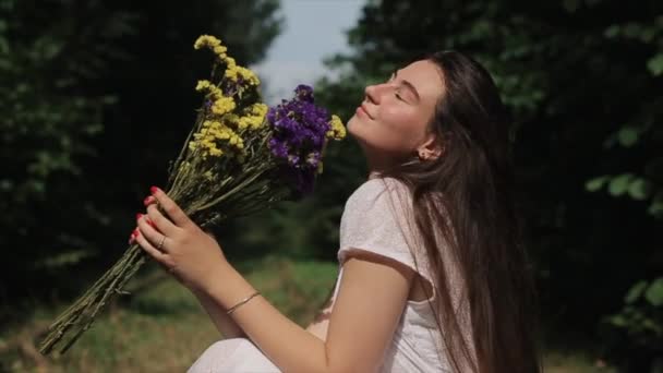 Egy fiatal mosolygós lány a zöld fák hátterében ül el, és arcát a nap felé fordítja, kezében egy csokor vadvirággal. Közelkép. Lassú mozgás. A kamera ráközelít. — Stock videók