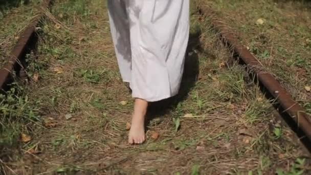 Una joven vestida de blanco largo camina descalza por un viejo ferrocarril abandonado en el bosque con un ramo de flores silvestres. Primer plano de las piernas. Movimiento lento — Vídeo de stock