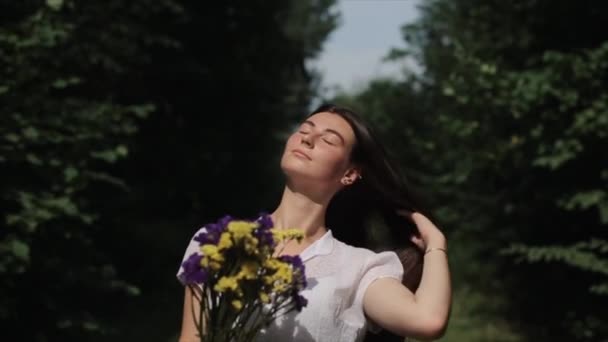 En öm ung kvinna i en vit klänning står med en bukett vilda blommor mot en bakgrund av träd och rätar ut hennes långa hår stänga ögonen. Närbild. Långsamma rörelser — Stockvideo