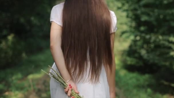 En öm ung kvinna i en vit klänning med mycket långt hår går mot bakgrunden av träd och vänder sig till kameran kastar blommor. Närbild. Utsikt bakifrån. Långsamma rörelser — Stockvideo
