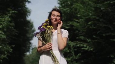 Beyaz elbiseli çekici genç bir kadın doğanın arka planında bir buket çiçekle duruyor ve kameraya bakarken gülümsüyor. Yakın plan. Ağır çekim. Düşük açı